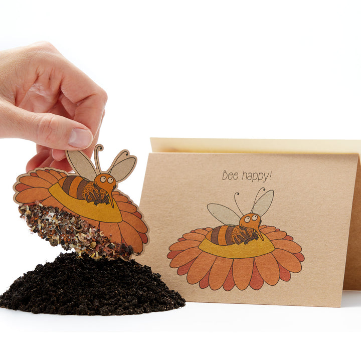 Eine Hand, die eine Aussaatkarte in Form einer Biene hochhält, mit dem Slogan "Bee happy!" darauf und einem Haufen Erde darunter.