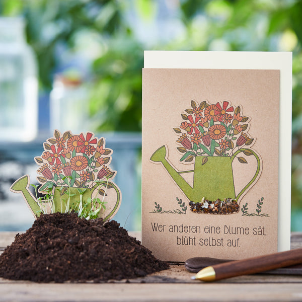 Grußkarte mit einer Illustration einer Gießkanne voller Blumen und dem deutschen Spruch „Wer anderen eine Blume sät, blüht selbst auf“, daneben eine Aussaatkarte im Blumentopf mit Erde und aufgehendem Grün.