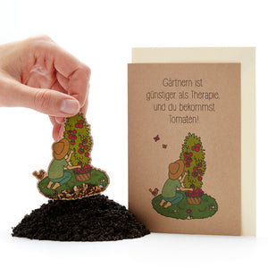 Eine Hand steckt eine Samenkarte mit einer Gärtnerin und einem Tomatenstrauch in Erde, daneben eine Grußkarte mit der Aufschrift „Gärtnern ist günstiger als Therapie, und du bekommst Tomaten!“