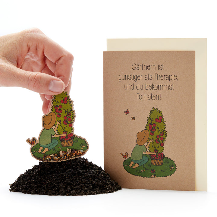 Eine Hand steckt eine Samenkarte mit einer Gärtnerin und einem Tomatenstrauch in Erde, daneben eine Grußkarte mit der Aufschrift „Gärtnern ist günstiger als Therapie, und du bekommst Tomaten!“