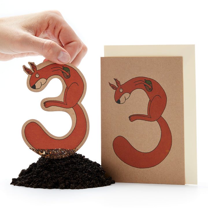 Hand hält eine rote Eichhörnchen-Pflanzfigur neben einer Grußkarte mit gleichem Design auf weißem Hintergrund.