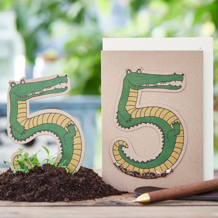 Kreative Darstellung von Zahlen als Krokodile auf Papier, neben Erde und Bleistift.