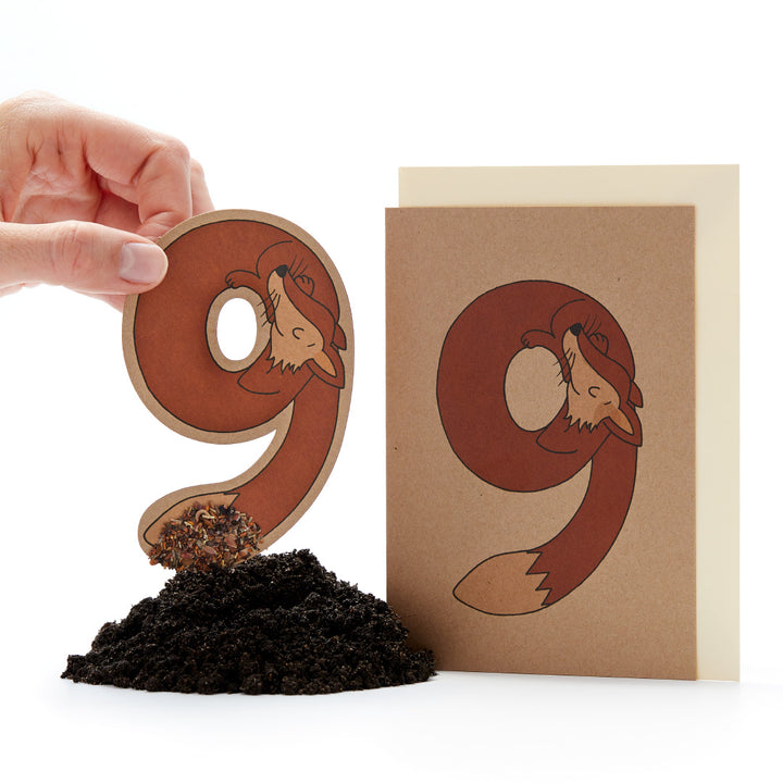 Hand hält einen ausgestanzten Buchstaben "ö" mit einem Fuchsdesign neben einer Karte mit dem gleichen Design und einem kleinen Erdhaufen.