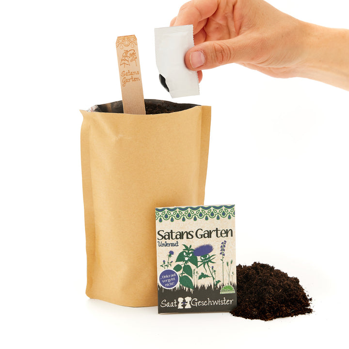 Eine Hand, die einen Samenbeutel über eine Papiertüte mit Erde hält, neben der ein Schild mit der Aufschrift "Satans Garten" und ein weiterer Samenbeutel stehen.