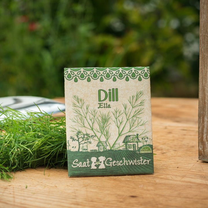 Päckchen Dillsamen mit der Aufschrift "Ella" von Saat-Geschwister auf einem Holztisch mit frischem Dill im Hintergrund.