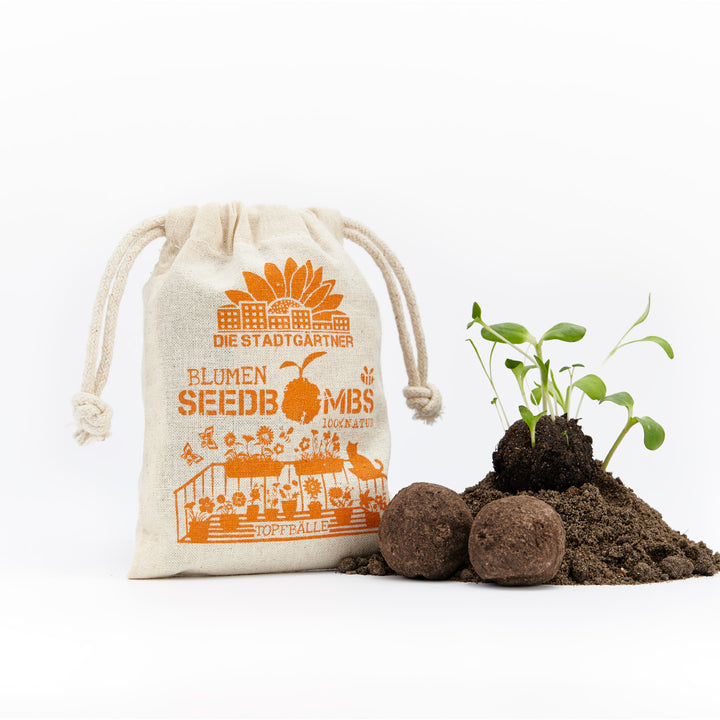 Stoffbeutel mit Aufschrift "DIE STADTGÄRTNER BLUMEN SEEDBOMBS 100% NATUR" und daneben keimende Pflanzen auf einem Erdhaufen mit zwei Seedbombs.