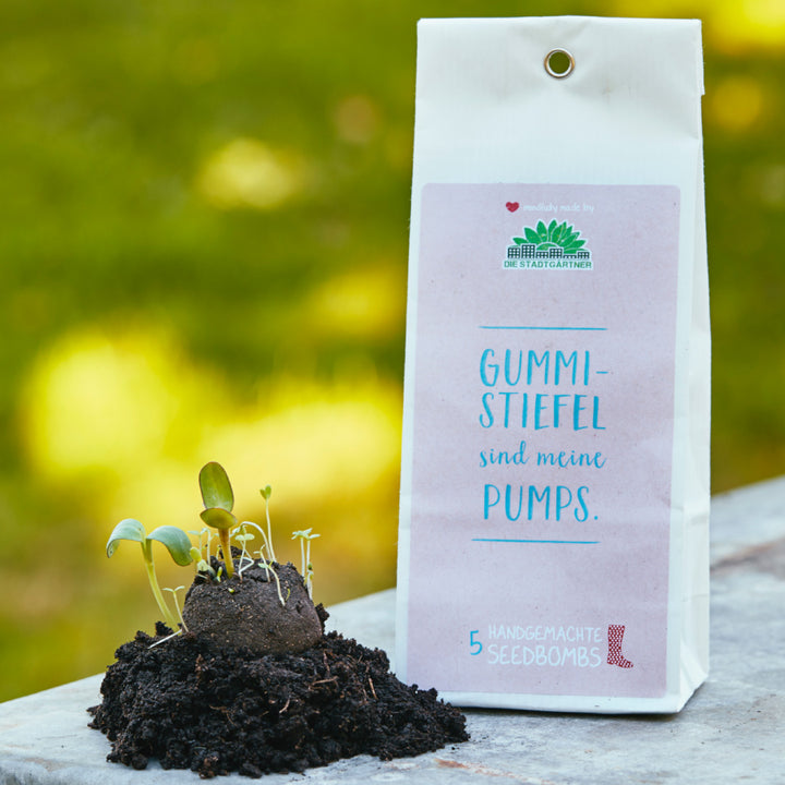 Verpackung von handgemachten Seedbombs mit dem Text "Gummi-Stiefel sind meine Pumps" und Keimlingen auf Erde daneben.