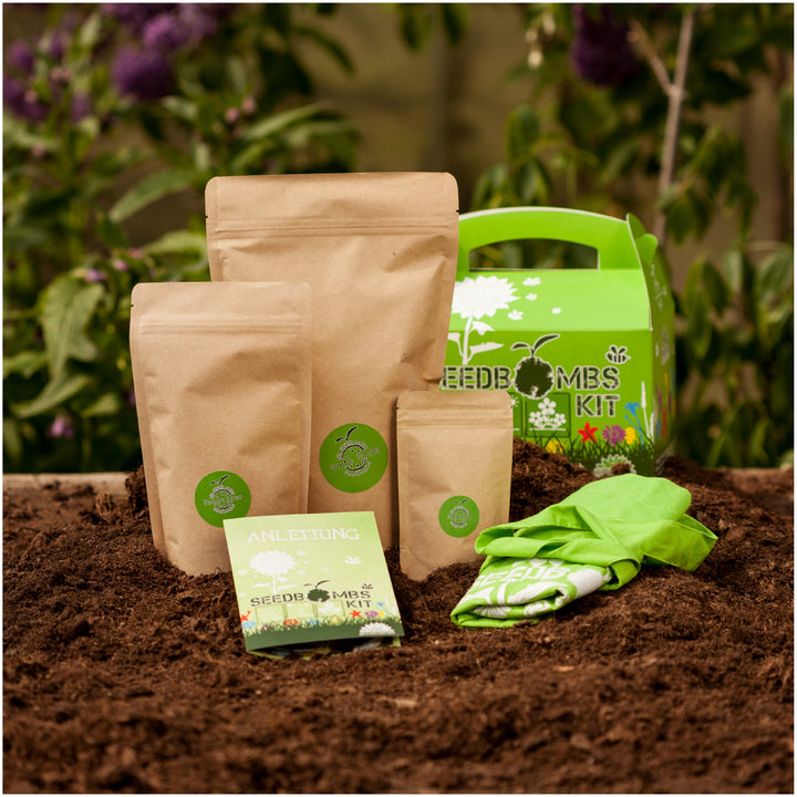 Gartenset mit Samenbomben, Verpackungen und Anleitung auf Erdboden, mit Pflanzen im Hintergrund.